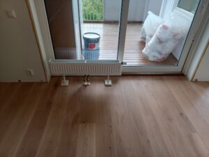 Ås - Bilde av et rom med nylagt parkettgulv, radiator under vinduet og malingsbøtte i bakgrunnen. Illustrerer prosessen med gulvlegging av en gulvlegger, med fokus på kvalitet og detaljer i gulvinstallasjonen.